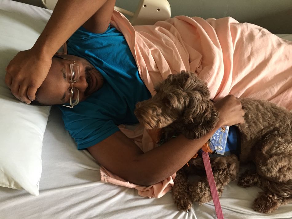 small brown dog lies alongside a patient in his bed. Patient has his arm around dog and is smiling.