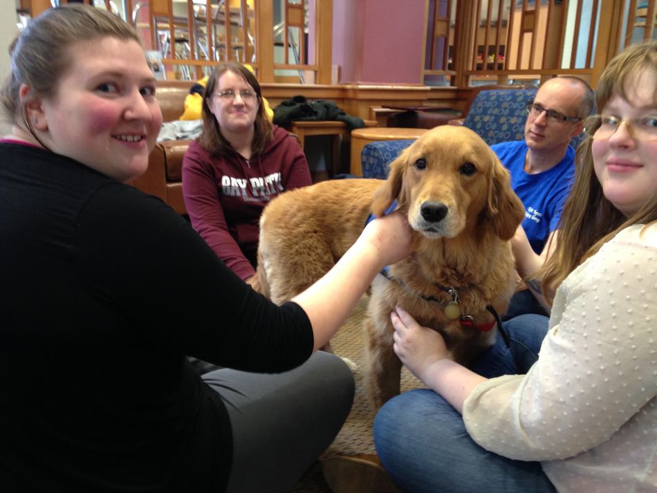 Golden Retriever sits encircled by students petting her and hugging her.