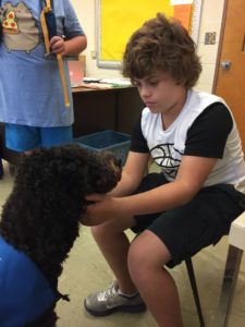 Betty sits in front of a male student sitting in a chair petting her.