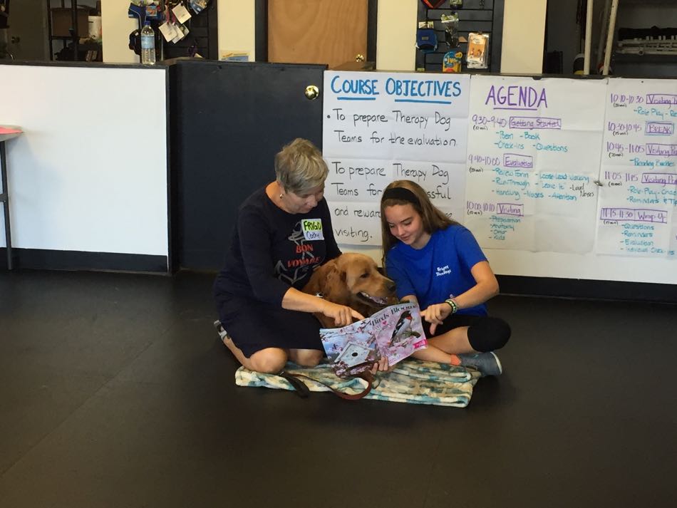 A woman, a dog, and a young girl sitting on the floor together looking at a book