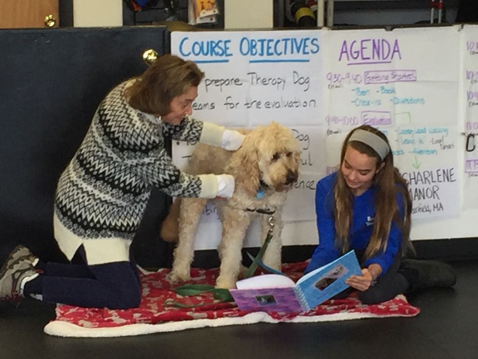 young girl sitting on floor reads from a book while a yellow dog and a women look on.